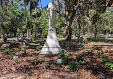 A white statue stands on a pedestal engraved with “The Spirit of Ecumenism” in a cemetery garden, surrounded by small plant markers on mulched ground.
