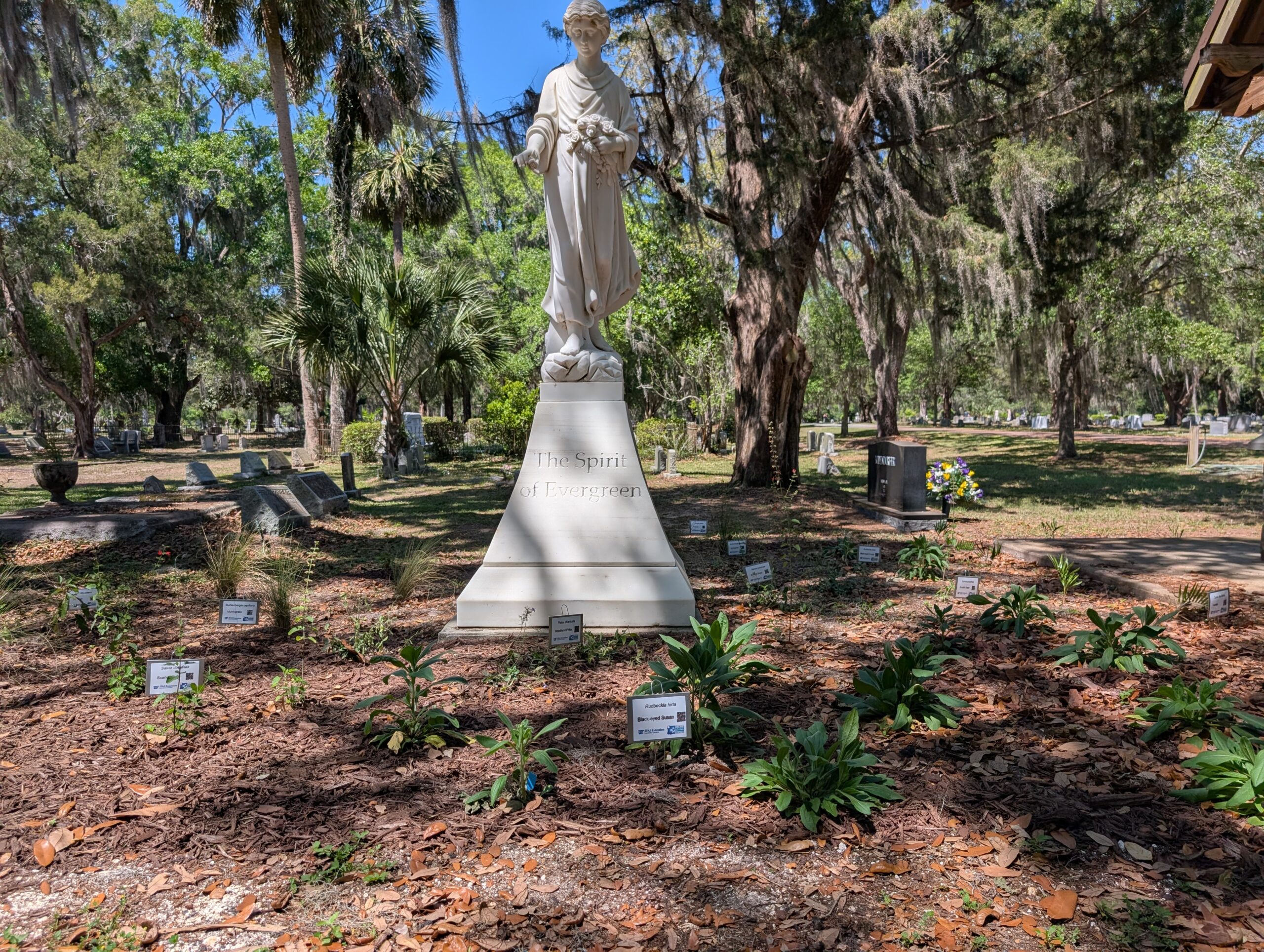 A white statue stands on a pedestal inscribed with The Spirit of Ecumenism, surrounded by small plants and labeled signs in a cemetery with trees in the background.