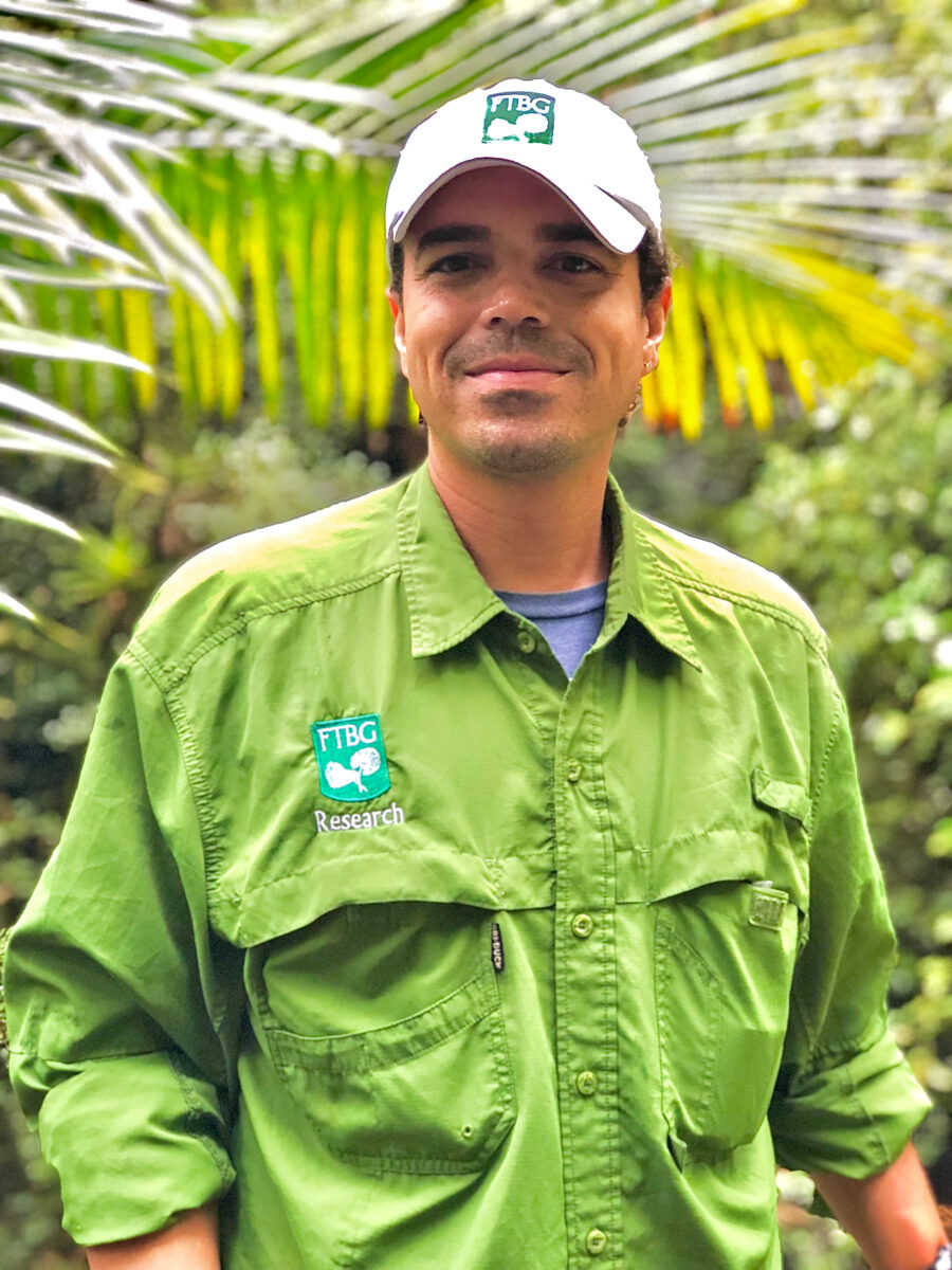 A person in a green FTBG Research shirt and white cap stands outdoors with tropical plants in the background.