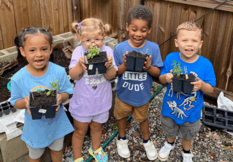 Four young children stand outdoors holding small potted plants, smiling at the camera, with a wooden fence and gardening supplies in the background.