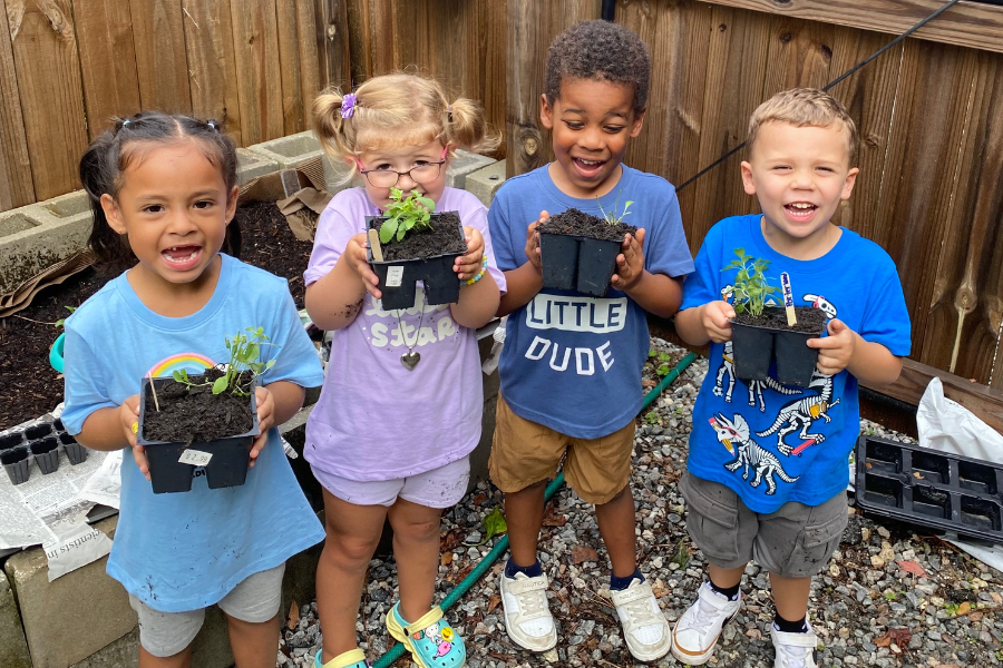 Four young children stand outdoors holding small potted plants, smiling at the camera, with a wooden fence and gardening supplies in the background.