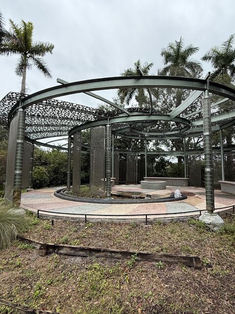 Open circular metal pavilion structure with decorative panels, situated outdoors amid palm trees and cloudy skies, surrounded by landscaping and pathways.