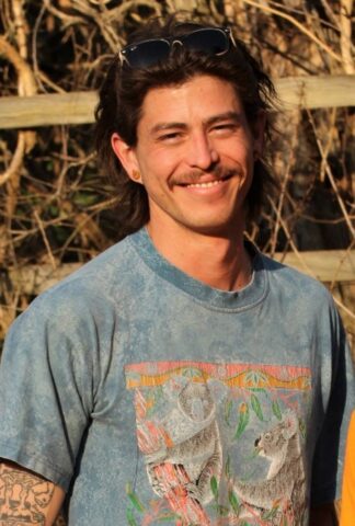 A man with long brown hair and a mustache smiles outdoors, wearing sunglasses on his head and a blue T-shirt with a koala design. Dry branches and a wooden fence are in the background.