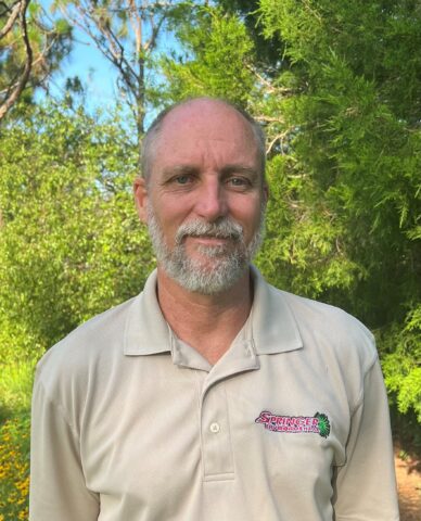 A man with a gray beard wearing a beige collared shirt stands outdoors in front of green trees and foliage.