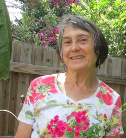 An older woman with short gray hair stands outside in a garden, smiling and wearing a white shirt with a red and green floral pattern. A wooden fence and lush plants are in the background.