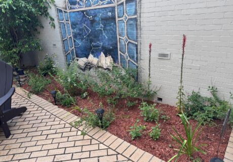 A small garden with green plants and mulch sits by a white brick wall featuring a mural of books and a blue stained glass window. Patio chairs and pathway lights are visible.