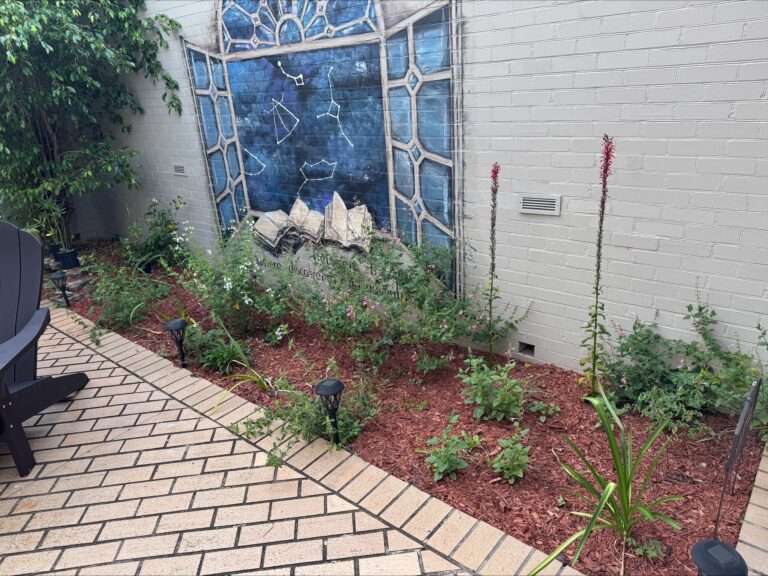A small garden with green plants and mulch sits by a white brick wall featuring a mural of books and a blue stained glass window. Patio chairs and pathway lights are visible.