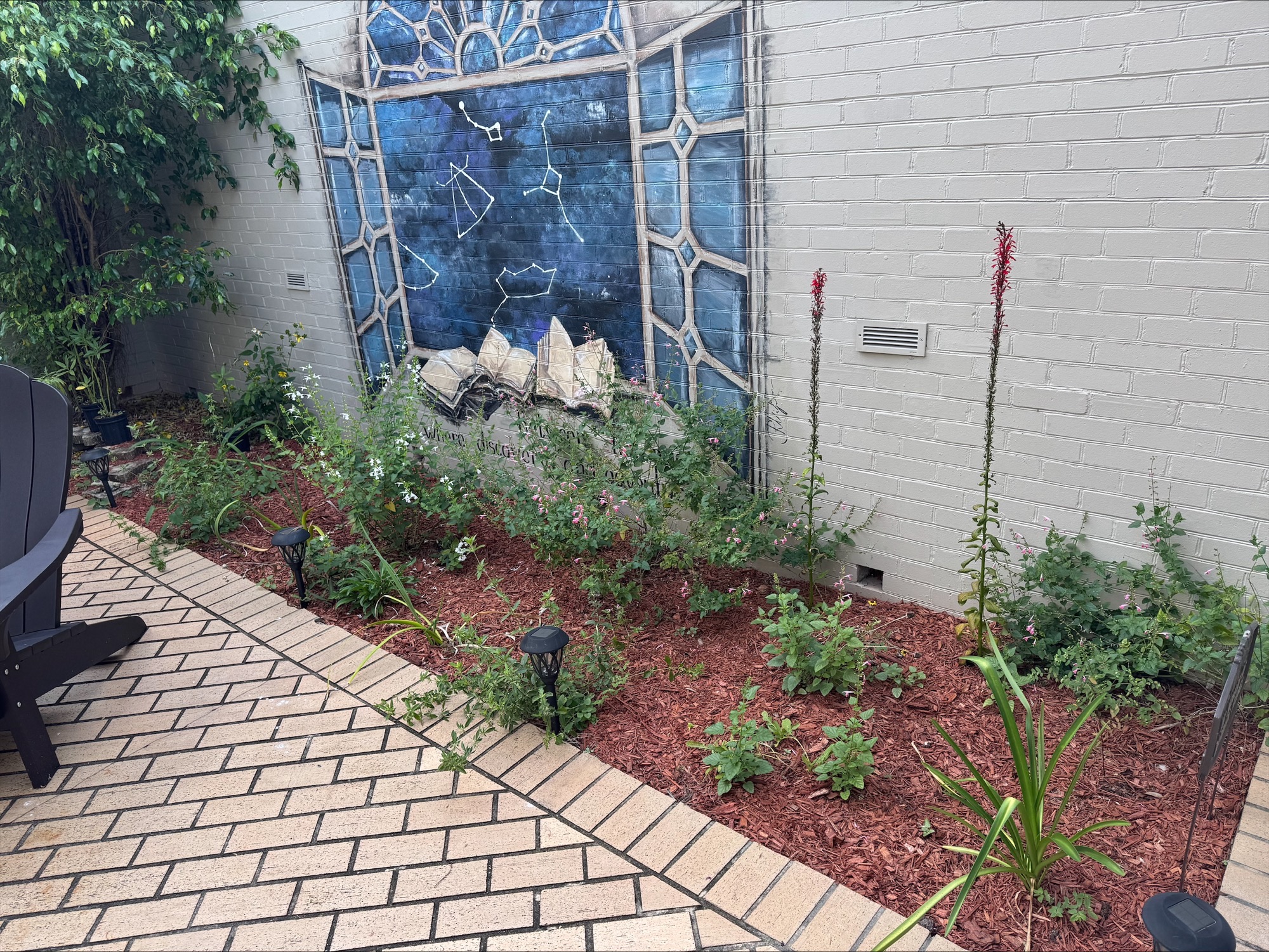 A small garden with green plants and mulch sits by a white brick wall featuring a mural of books and a blue stained glass window. Patio chairs and pathway lights are visible.