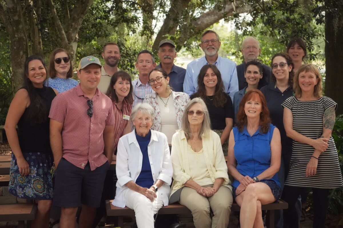 A group of seventeen people, including men and women of various ages, pose for a photo outdoors on wooden benches in front of trees and greenery.
