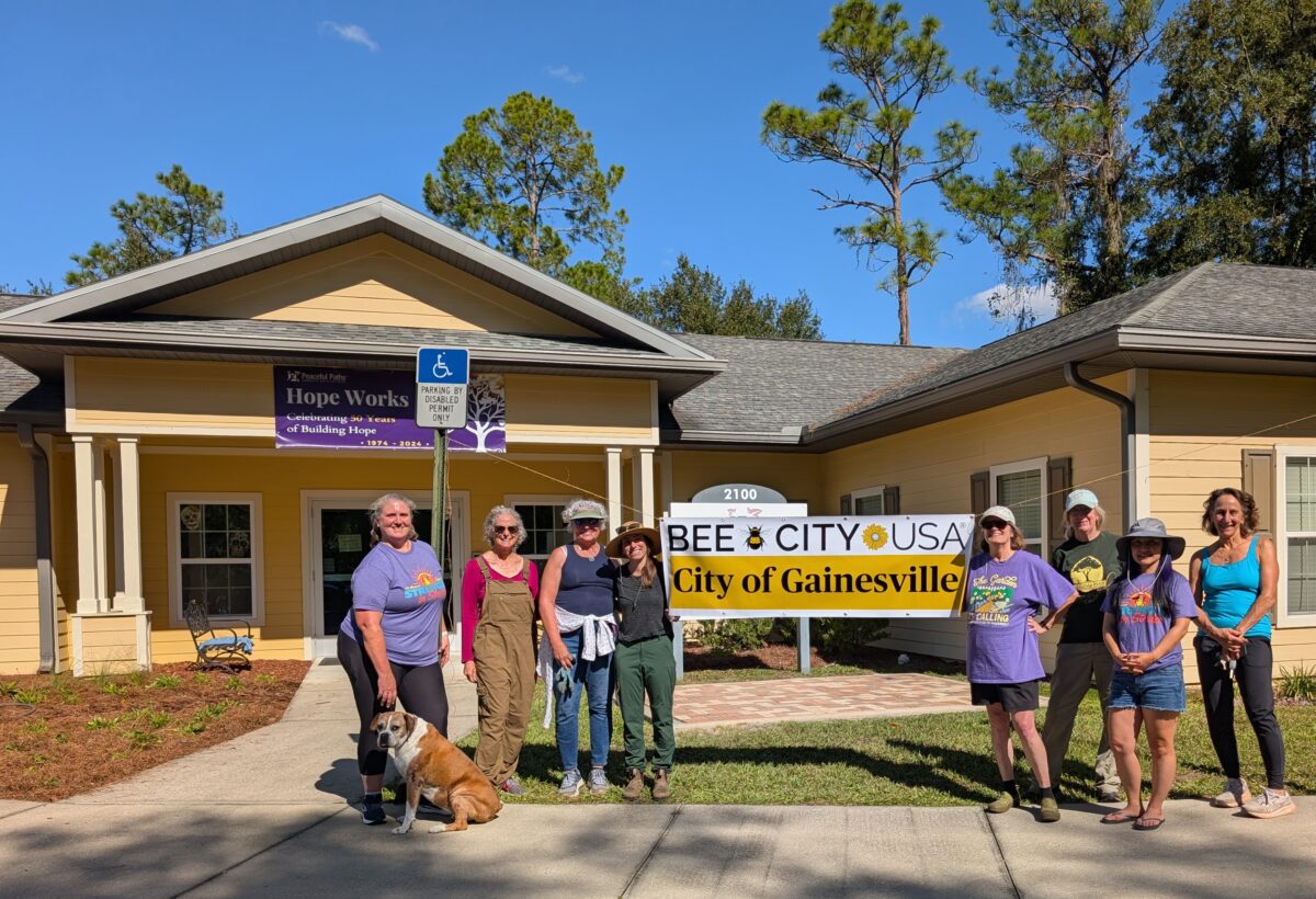 A group of people and a dog stand outside a yellow building with a sign that reads BEE CITY USA City of Gainesville.