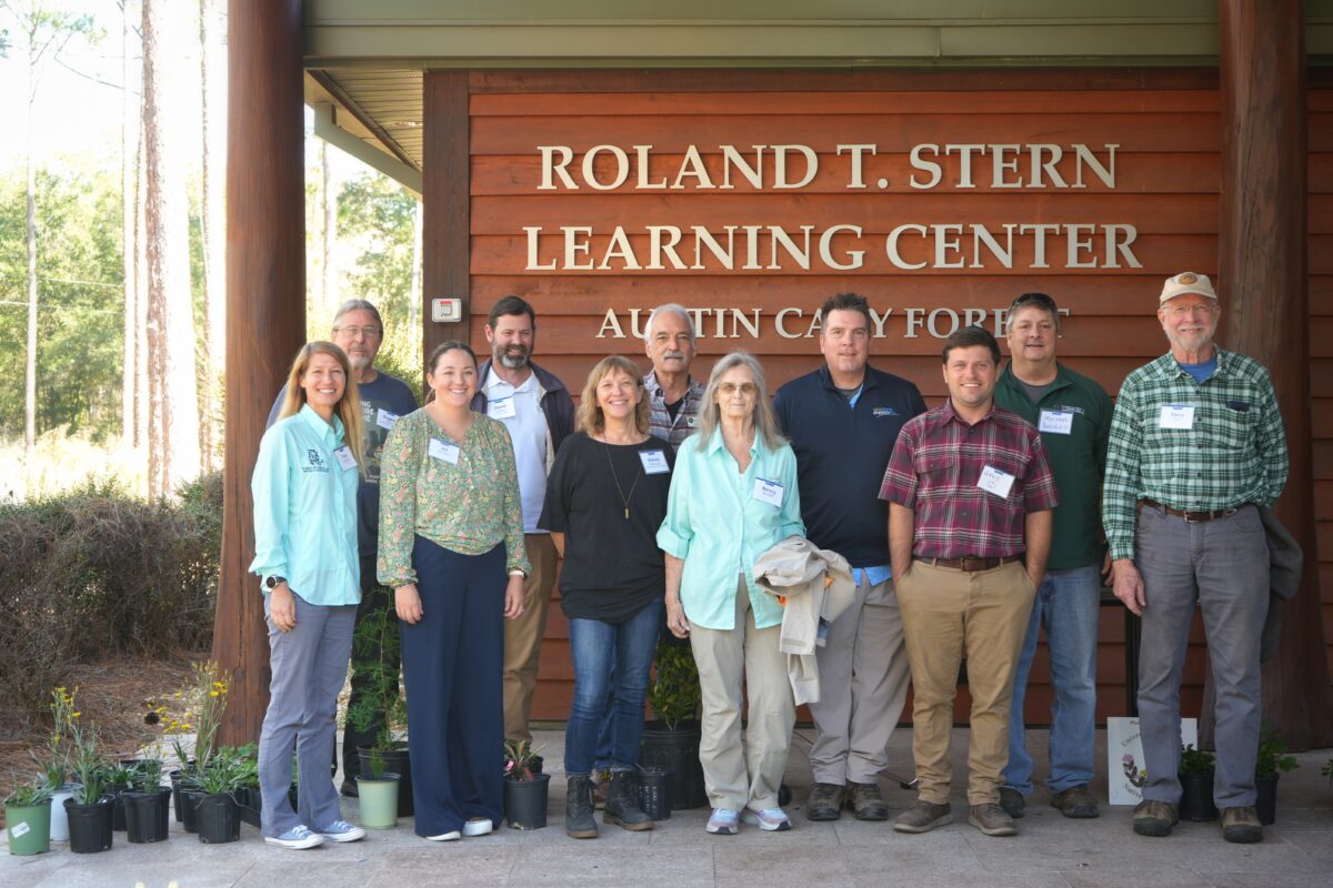 Twelve people stand in front of the Roland T. Stern Learning Center, with plants in pots on the ground near them.
