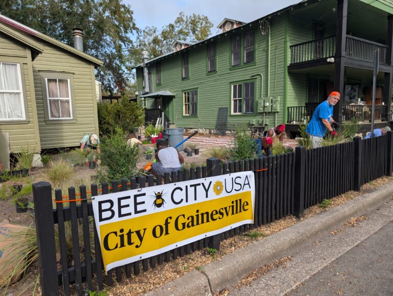 People are gardening in front of a green house behind a black fence with a Bee City USA - City of Gainesville sign attached to it. Decorations and gardening tools are visible.