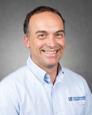 A man with short brown hair smiles at the camera, wearing a light blue UF IFAS Extension University of Florida button-up shirt, against a plain gray background.