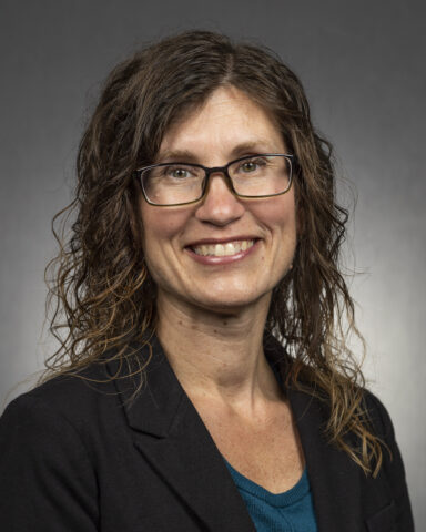 A woman with wavy brown hair, glasses, and a black blazer over a teal top, smiling at the camera against a plain gray background.