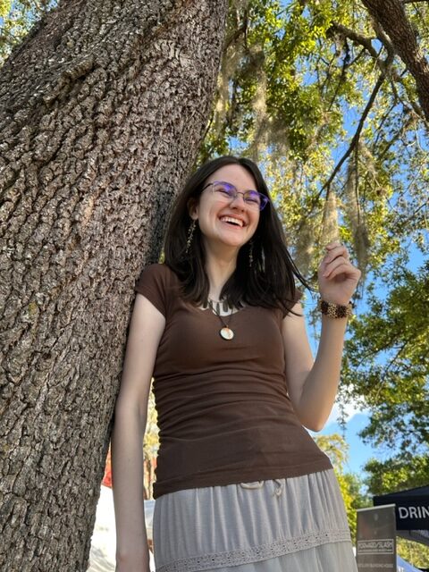 A young woman wearing glasses and a brown top stands smiling next to a large tree on a sunny day, with green leaves and blue sky in the background.