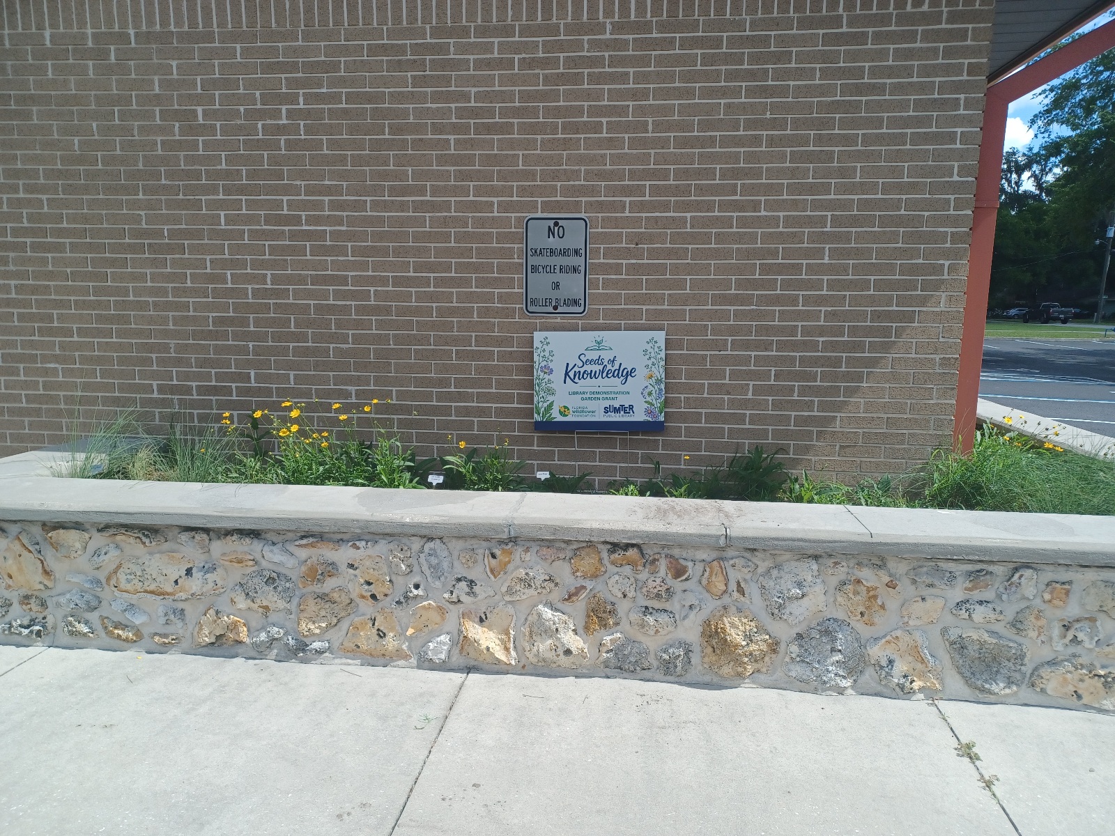 A brick wall with a No skateboarding or bicycling sign and a Seeds of Knowledge garden sign behind a stone-edged flower bed with various plants.