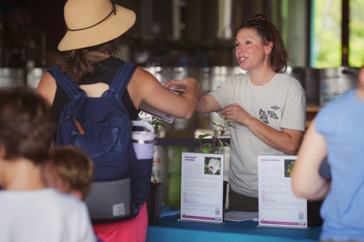 A woman behind a table hands a plant to another woman with a backpack. Informational sheets and potted plants are displayed on the table.