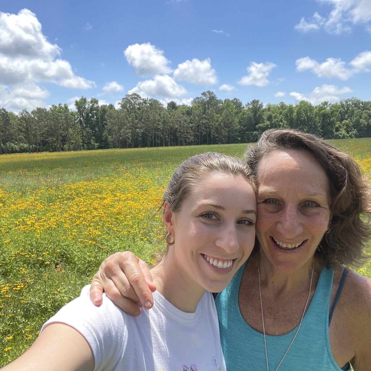 Two women smiling and taking a selfie together in front of a field of yellow wildflowers with trees and a blue sky in the background.