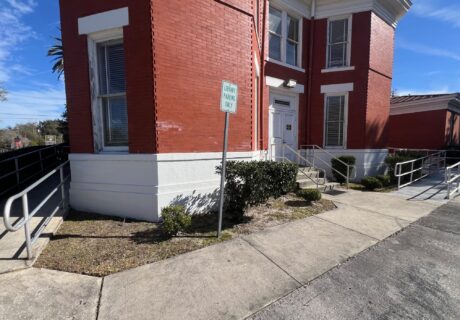 Red brick building with white trim, a sidewalk, multiple ramp railings, a small sign reading Library Parking Only, and some shrubs near the entrance.