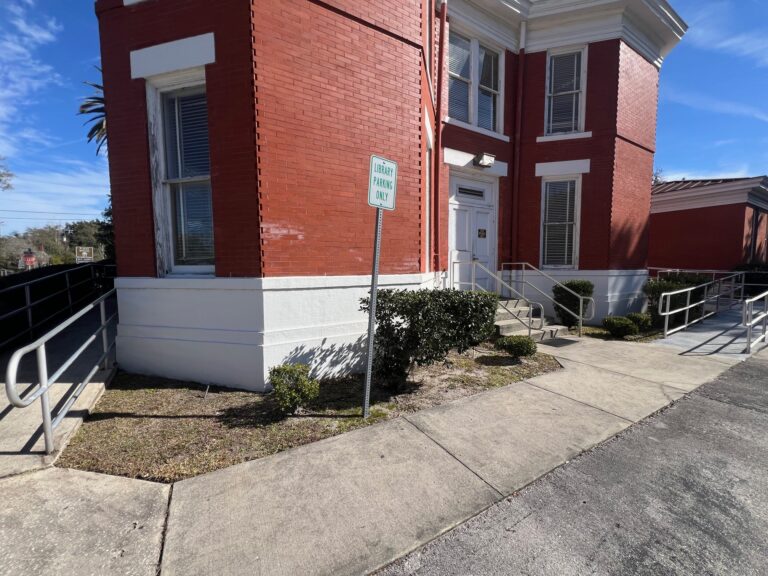 Red brick building with white trim, a sidewalk, multiple ramp railings, a small sign reading Library Parking Only, and some shrubs near the entrance.