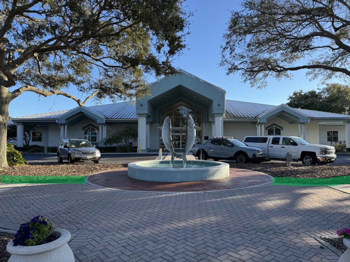 Single-story building with a metal roof, cars parked in front, and a circular fountain with fish sculptures at the entrance, surrounded by trees and landscaping.
