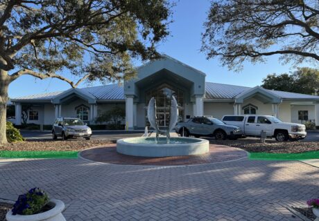 Single-story building with a metal roof, cars parked in front, and a circular fountain with fish sculptures at the entrance, surrounded by trees and landscaping.