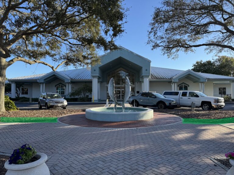 Single-story building with a metal roof, cars parked in front, and a circular fountain with fish sculptures at the entrance, surrounded by trees and landscaping.