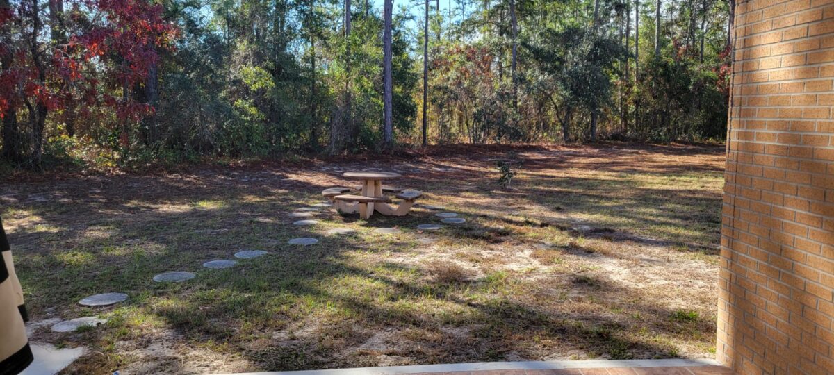 A small round picnic table with benches sits on a grassy area with stepping stones, surrounded by trees and bordered by a brick wall on the right.