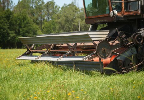 A close-up of a combine harvester cutting through a grassy field with trees in the background.