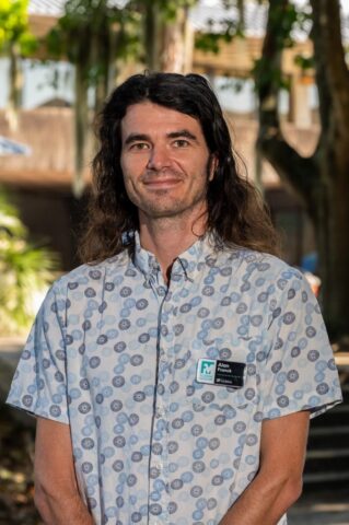 A man with long brown hair, wearing a patterned short-sleeve shirt and a name badge, stands outdoors in front of trees and a building.