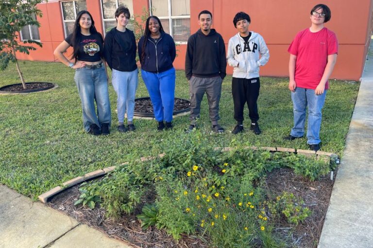 Six people stand behind a small garden bed with yellow flowers, outside near a building with red-orange walls.