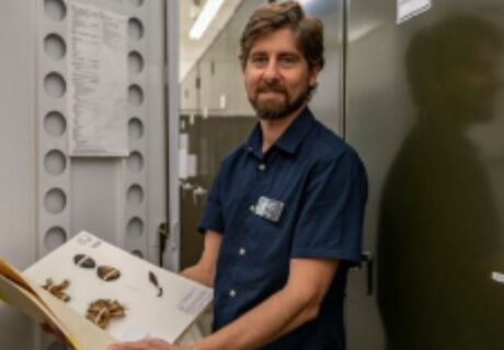 A man with a beard stands in a storage room holding an open folder displaying preserved plant specimens.