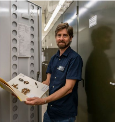 A man stands in a storage room holding an open book displaying insect specimens, with metal cabinets in the background.