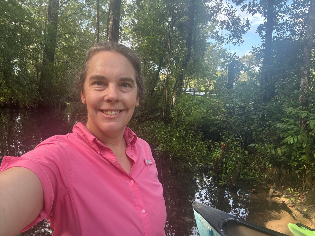 A person in a pink shirt takes a selfie outdoors near a wooded area with water and a kayak visible in the foreground.