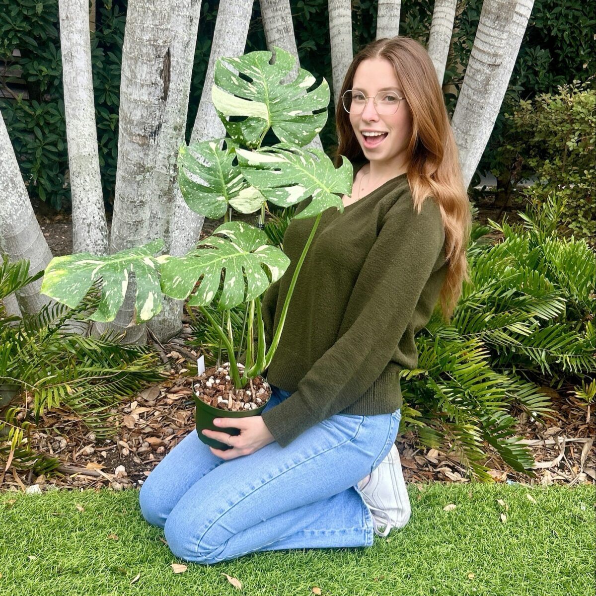 A woman with long hair, wearing glasses, a green sweater, and jeans, kneels on grass holding a potted monstera plant in an outdoor garden with trees and shrubs.