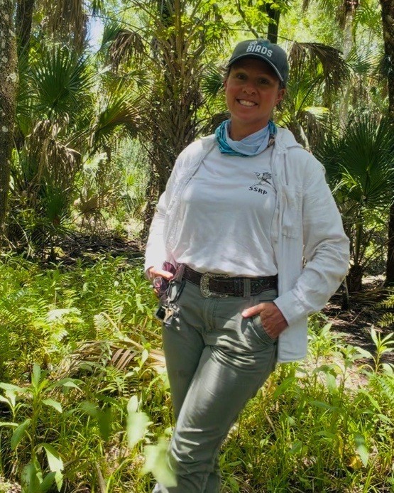 Person in outdoor gear stands in a sunny, forested area with palm trees and green plants, smiling at the camera.