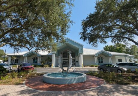 A building with a light blue roof and a fish fountain in front, surrounded by trees and parked cars on a sunny day.
