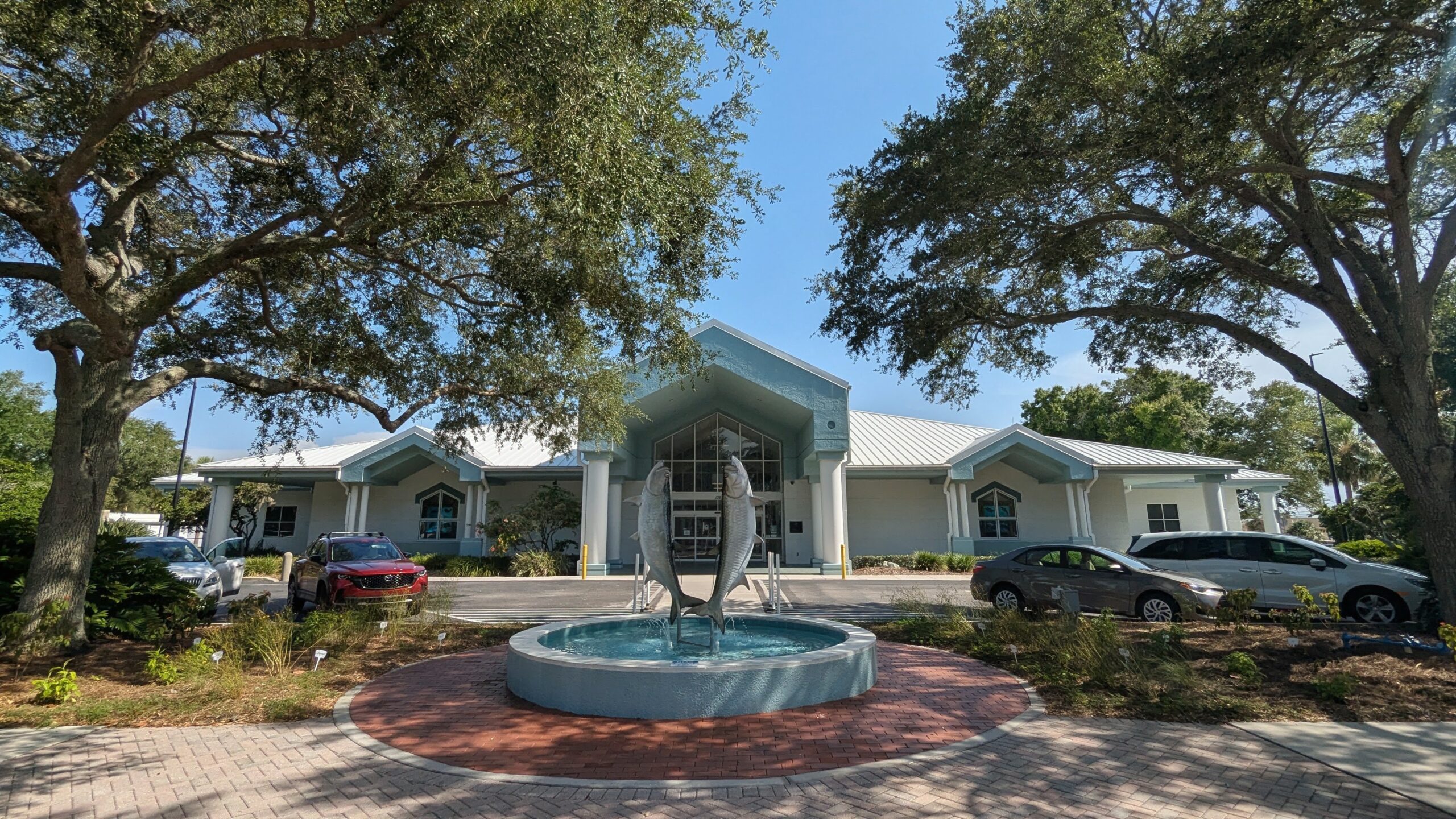 A building with a light blue roof and a fish fountain in front, surrounded by trees and parked cars on a sunny day.