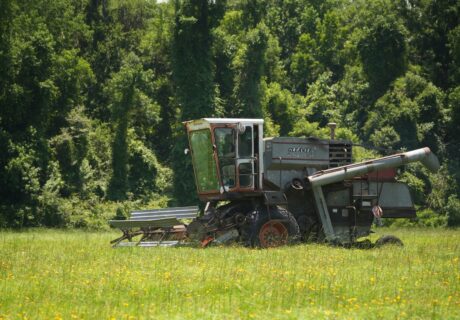 A gray combine harvester sits idle in a grassy field with dense, green trees in the background.