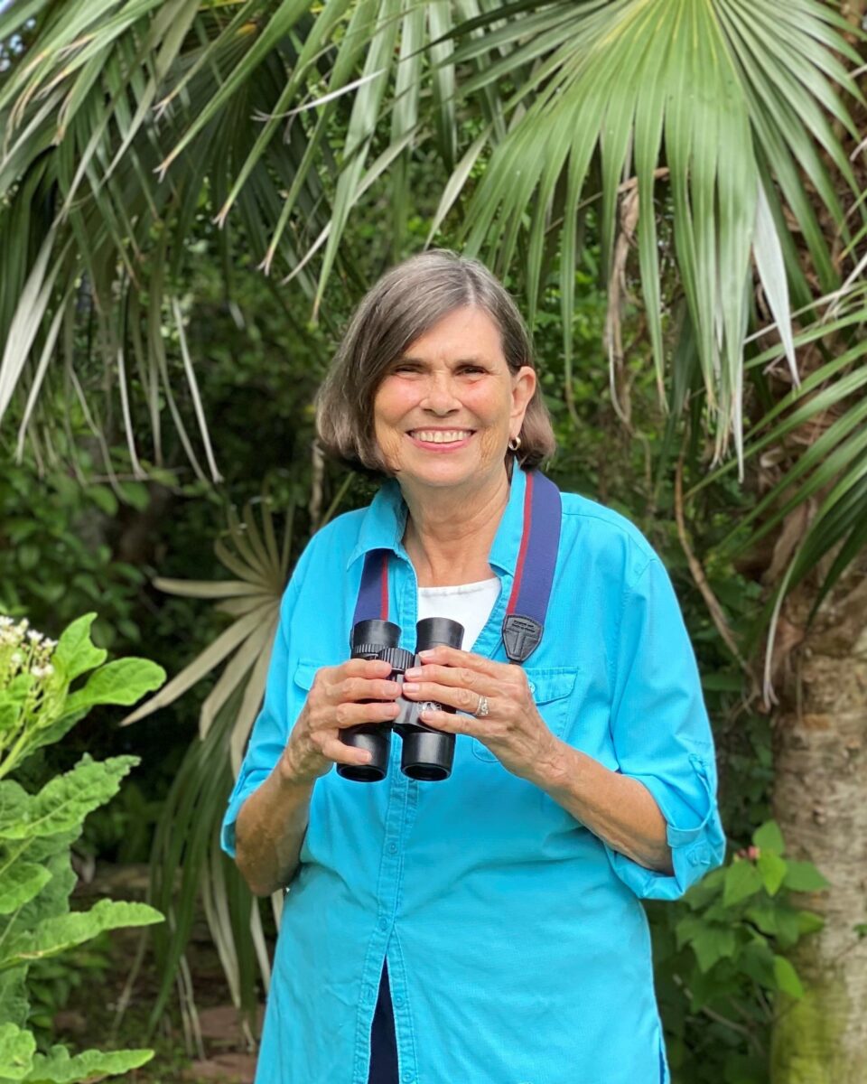 Older woman in a blue shirt stands outdoors under palm leaves, smiling and holding binoculars with both hands.