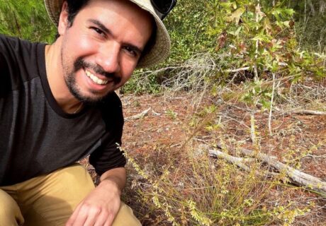 A person wearing a hat and dark shirt smiles while kneeling outdoors next to small green plants and dry branches on the ground.
