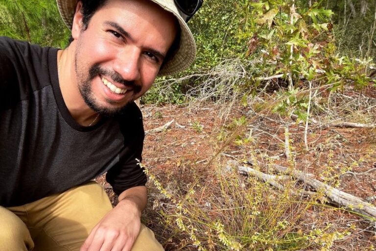 A person wearing a hat and dark shirt smiles while kneeling outdoors next to small green plants and dry branches on the ground.