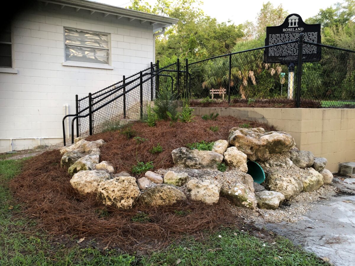 A sloped garden with rocks, mulch, a green drainage pipe, and a historical marker near a white building and black fence.