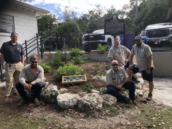 Five men pose around a small, landscaped garden bordered with rocks, with a sign in front and vehicles parked behind a black fence in the background.